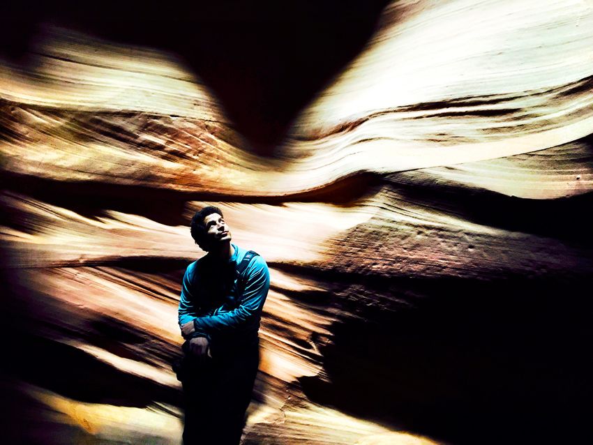 A man with dark hair wears a blue shirt with his arms crossed as he leans against the wall of a slot canyon.